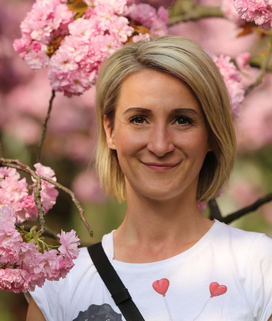 Tiersitterin lächelt auf Profilbild vor blühenden Kirschbäumen, trägt ein T-Shirt mit Herzmotiv.